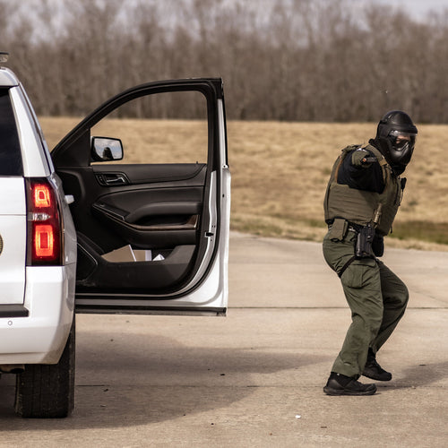 Person in tactical gear standing next to a police vehicle with open door.